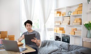 Seorang pria duduk di meja kerja di rumah sambil memegang clipboard, dengan laptop terbuka dan rak berisi kotak paket di belakangnya, menunjukkan aktivitas bisnis online atau pengelolaan pesanan dari rumah.
