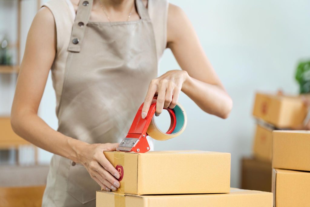 Seseorang mengenakan apron sedang menutup kotak kardus menggunakan lakban dengan alat dispenser, menunjukkan proses pengemasan paket untuk pengiriman.