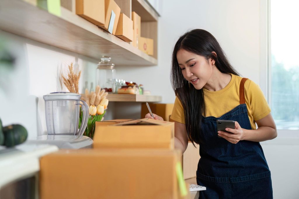 Seorang wanita mengenakan apron menulis detail pada kotak paket sambil memegang ponsel di dapur, menunjukkan aktivitas pengemasan dan pencatatan pesanan bisnis rumahan.