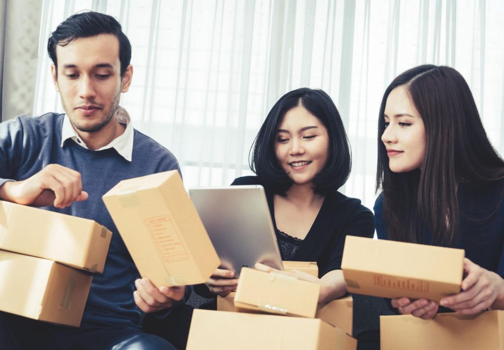 Tiga orang sedang memeriksa kotak paket bersama, dengan satu orang melihat tablet dan lainnya memegang kardus, menunjukkan aktivitas pengecekan pesanan atau proses pengemasan dalam tim.