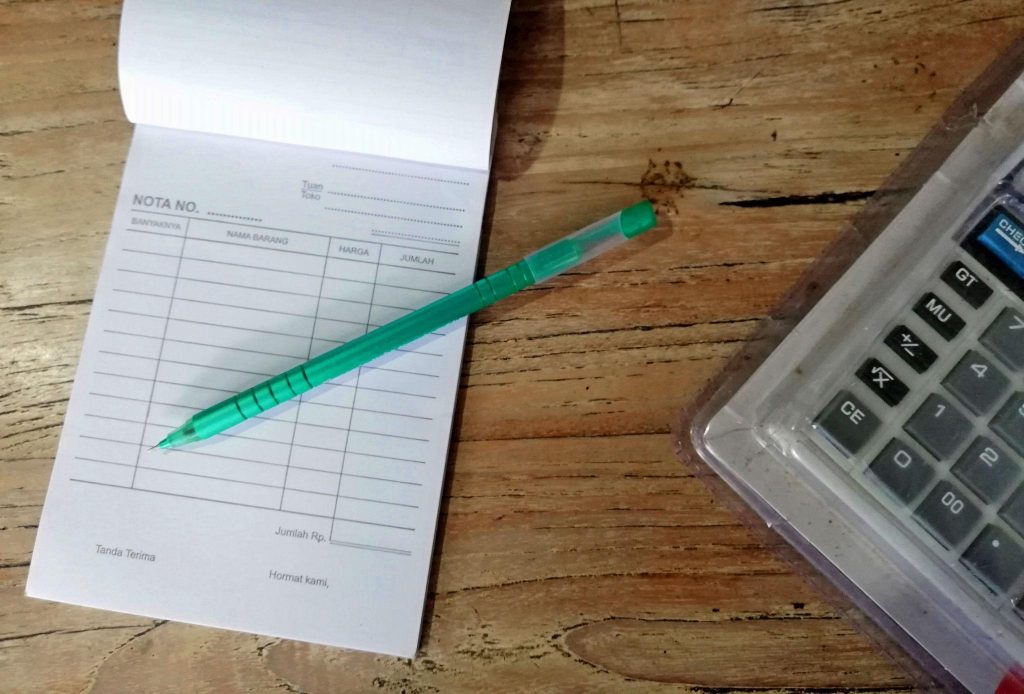 Buku nota, pulpen, dan kalkulator di atas meja kayu yang menggambarkan proses menghitung estimasi modal bisnis supplier.