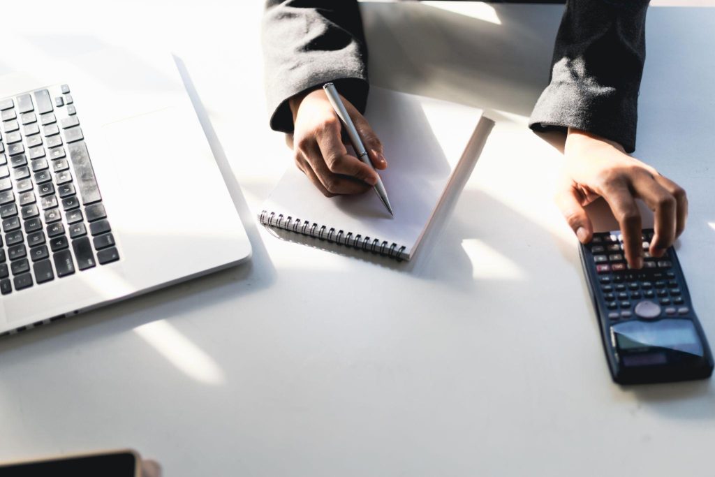 Seseorang mencatat anggaran sambil menggunakan kalkulator dan laptop yang menggambarkan perencanaan biaya pemasaran dalam bisnis supplier.