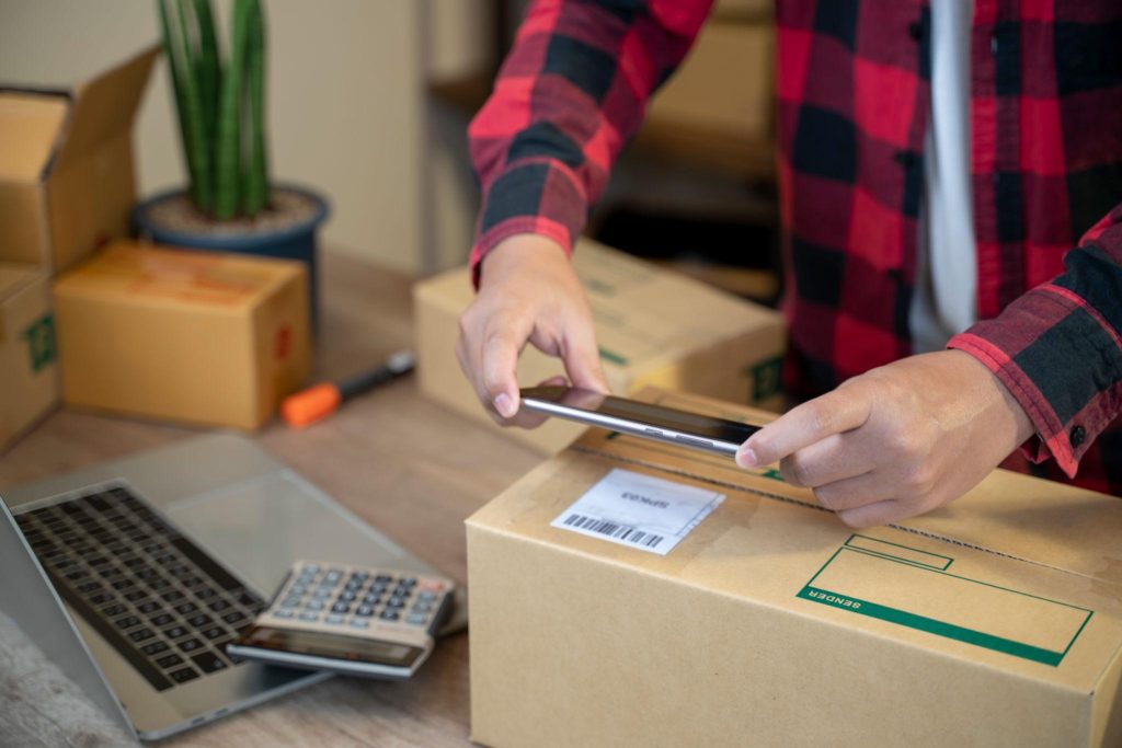 Seseorang menggunakan smartphone untuk mengelola paket barang dengan laptop dan kalkulator di meja sebagai bagian dari infrastruktur bisnis supplier.