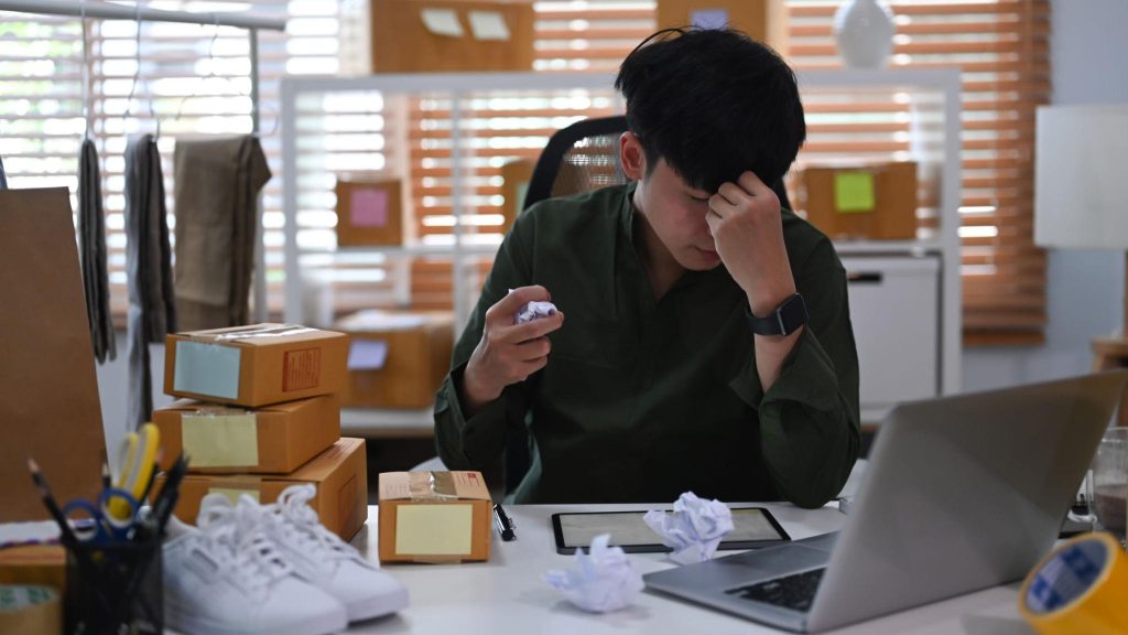 Seorang pelaku usaha terlihat stres di depan laptop dan paket barang